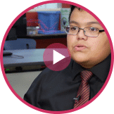 A person wearing glasses and a black shirt with a red tie is speaking. They are shown in a circular frame with a play button overlay. A desk and shelves in the blurred background hint at teaching entrepreneurship, possibly part of a K12 entrepreneur curriculum video.