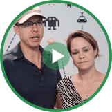 Two people stand and speak in front of a background with various icons, including a robot and other illustrations. The man wears a cap and eyeglasses, while the woman wears a patterned top and necklace, discussing the importance of youth entrepreneurship education.