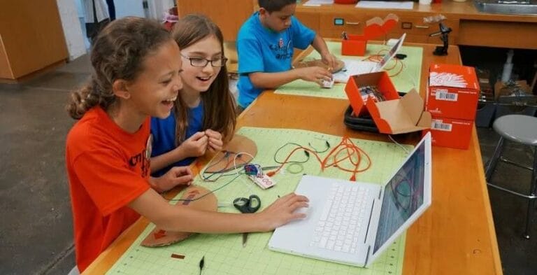 Three children are sitting at a table using laptops and various electronic components for a project. Two girls smile and interact with a laptop while a boy in the background focuses on his work, all part of an engaging youth entrepreneurship education program.
