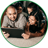 A family of four, consisting of two adults and two children, is lying on the floor looking at a tablet screen together, exploring a program on youth entrepreneurship education.