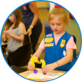A young girl in a blue uniform vest focuses on a craft project at a table, embodying the spirit of youth entrepreneurship education. Another child and an adult are in the background, also engaged in activities that foster creativity and initiative.