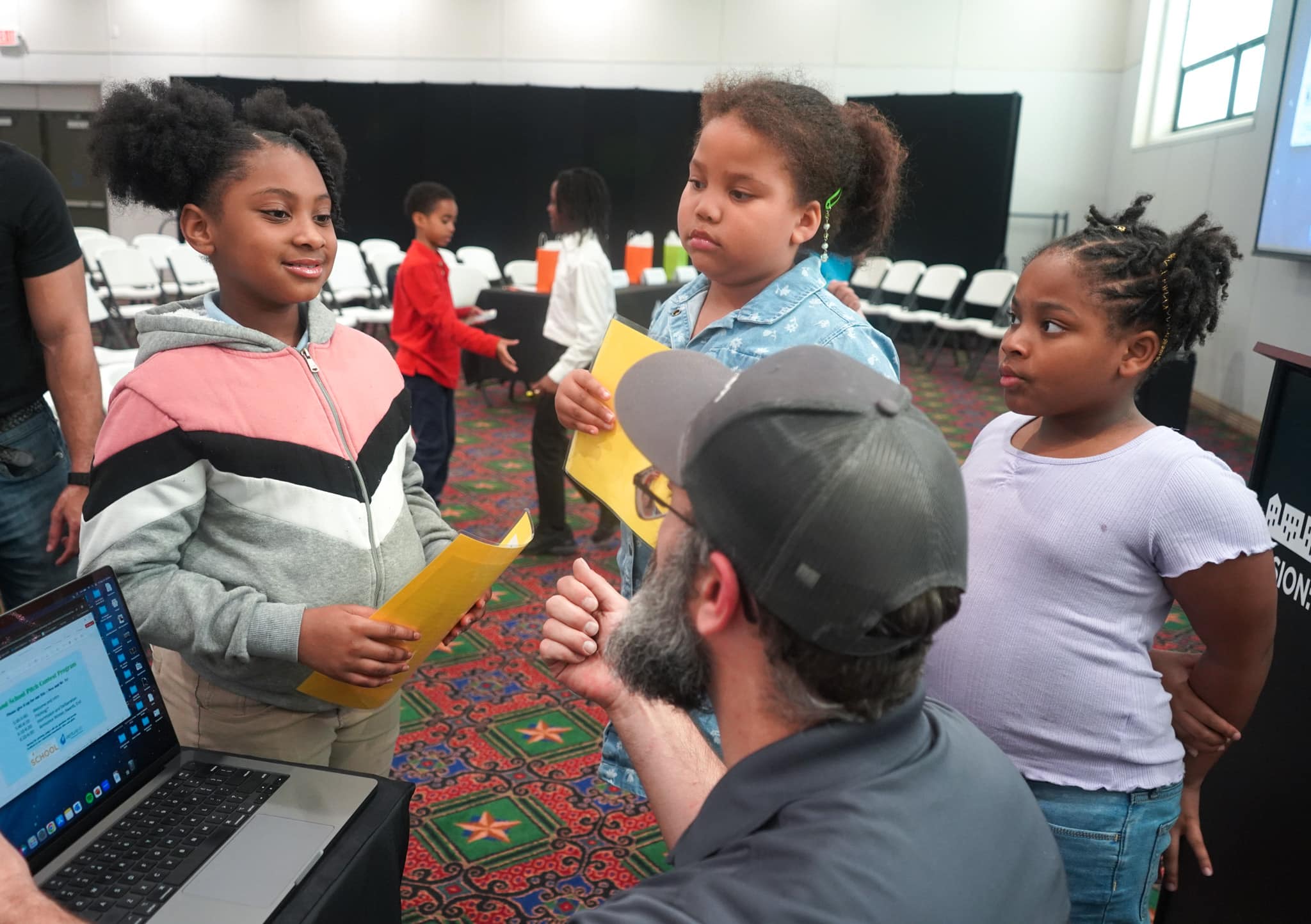 Children hold certificates while a man seated at a laptop appears to speak to them in a classroom setting, highlighting their achievements in the k-12 entrepreneurship curriculum.