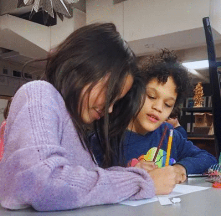 Two children are sitting together at a table, drawing on paper with pencils. One child is wearing a purple sweater, and the other has curly hair and a dark blue shirt. Their creativity is inspired by the spirit of VentureLab collaboration, fostering future innovators.